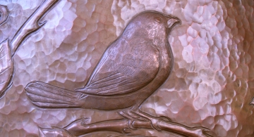 An exquisitely engraved Blackcapped-Chickadee in repoussé occupies a thicket of wild roses on a hammered copper vessel.